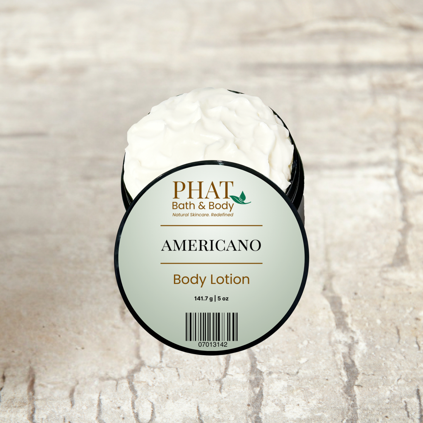 An open round tub of PHAT Bath & Body 'Americano' body lotion, showing the white cream inside, set against a light beige stone-textured background.