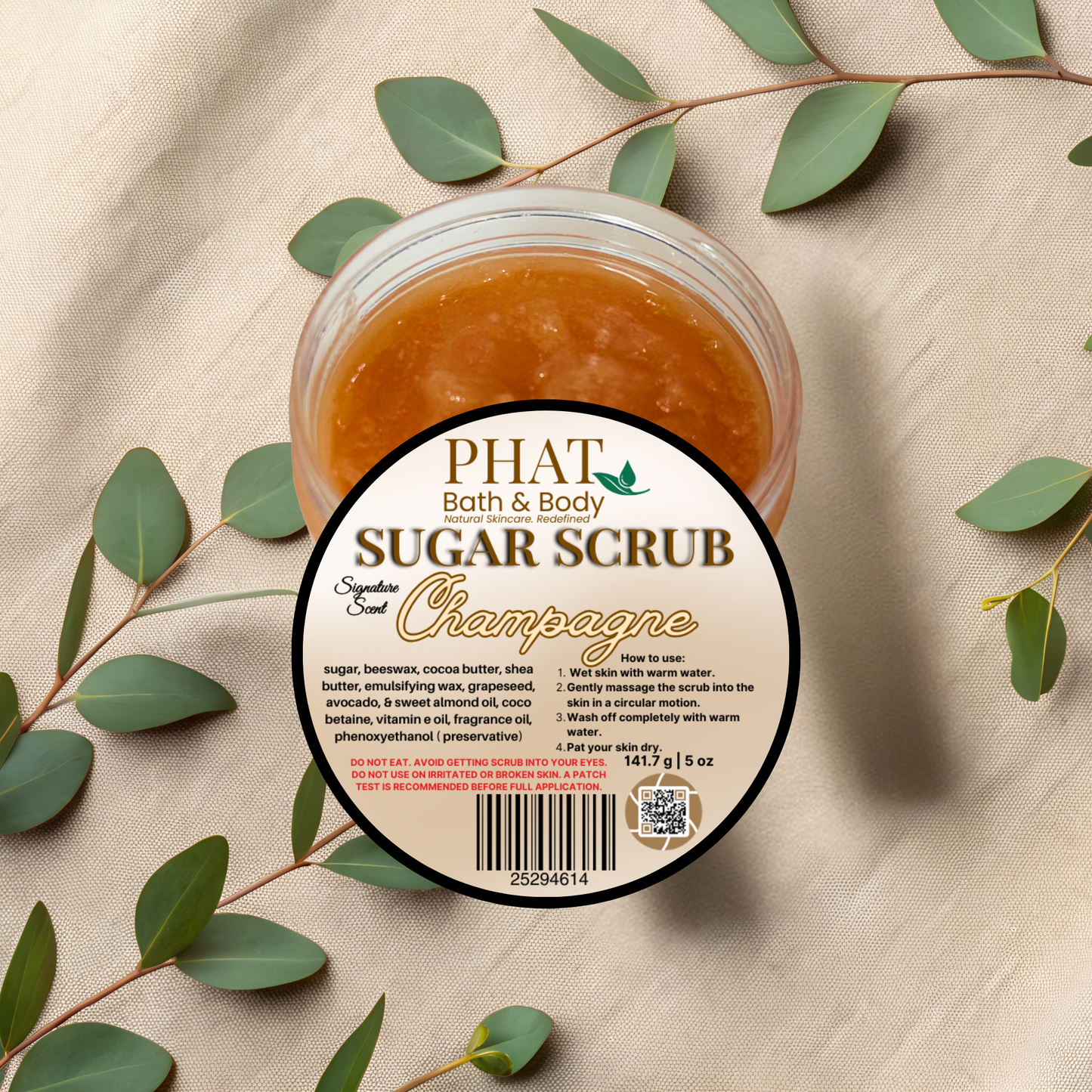 Jar of Phat Bath & Body Sugar Scrub with a 'Champagne' scent on a beige fabric background with green leaves.