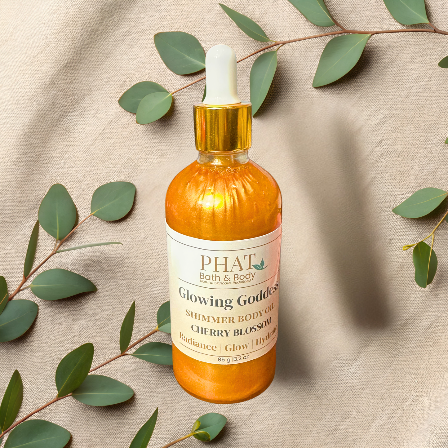 Bottle of PHAT Bath & Body Glowing Goddess Shimmer Body Oil on a beige fabric background with green leaves.