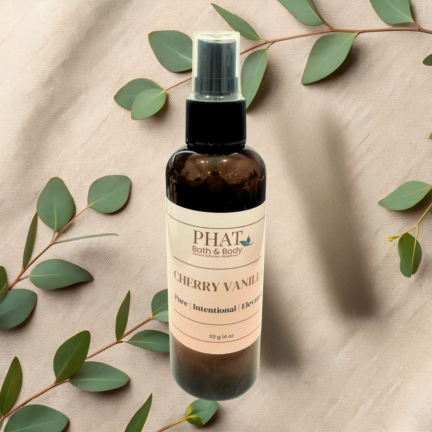Bottle of 'Cherry Vanilla' body spray from Phat Bath & Body on a beige fabric background with green leaves.