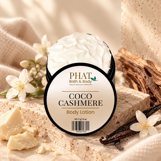 Phat Bath & Body Coco Cashmere 5 oz Body Lotion in an open black jar with labeled lid, styled with cocoa butter, vanilla beans, white florals, warm wood, and soft neutral fabric.
