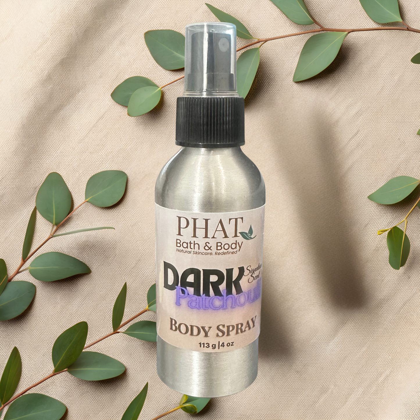 Body spray bottle labeled 'PHAT Bath & Body Dark Patchouli' on a beige fabric background with green leaves.