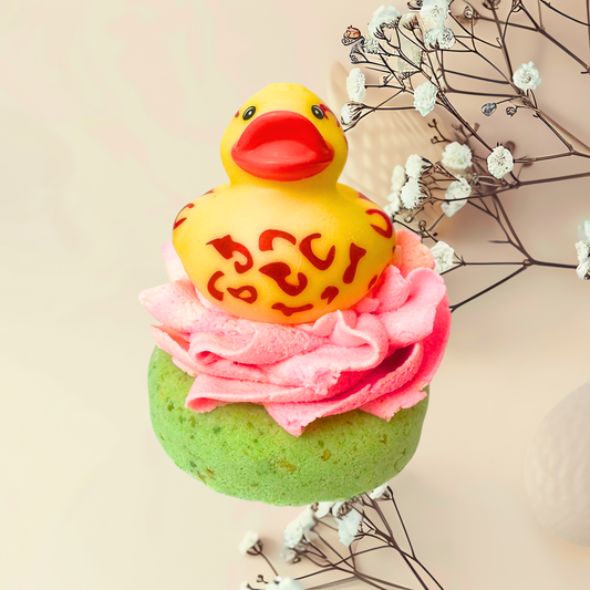 Yellow rubber duck on a green bath bomb with pink top, surrounded by white flowers and branches.