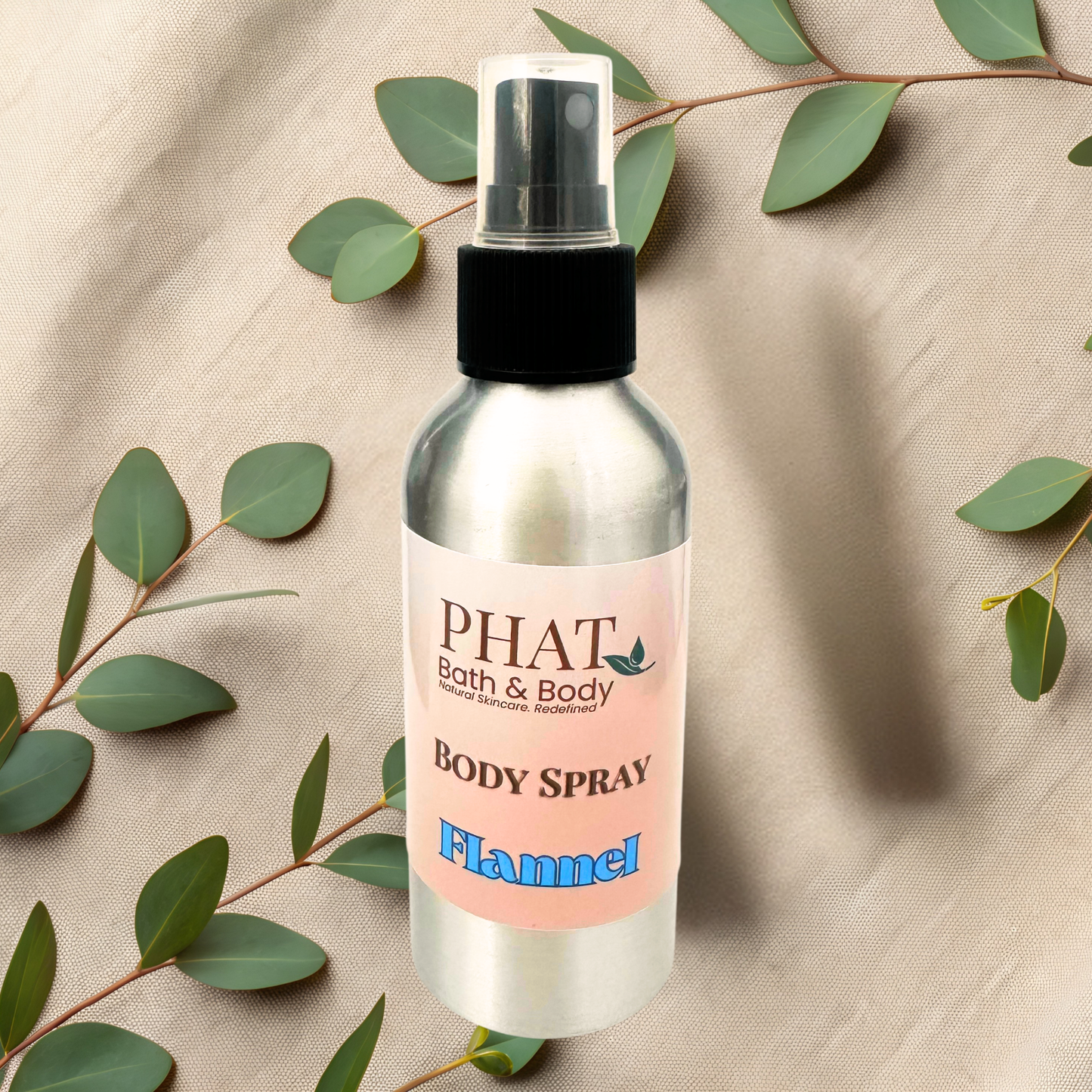 Body spray bottle labeled 'PHAT Bath & Body' with 'Flannel' scent on a beige fabric background with green leaves.