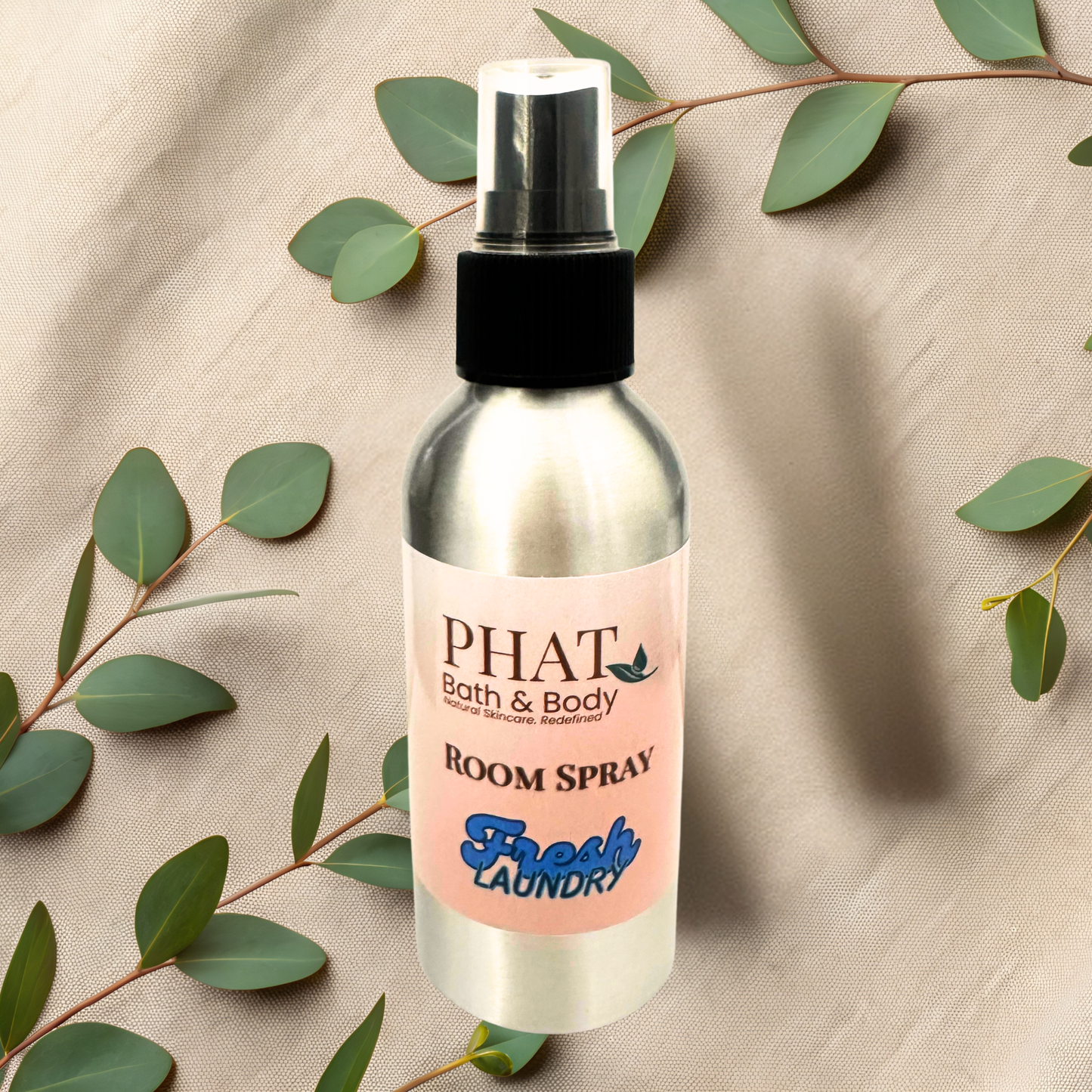 Bottle of PHAT Bath & Body room spray with a pink label on a beige fabric background with green leaves.