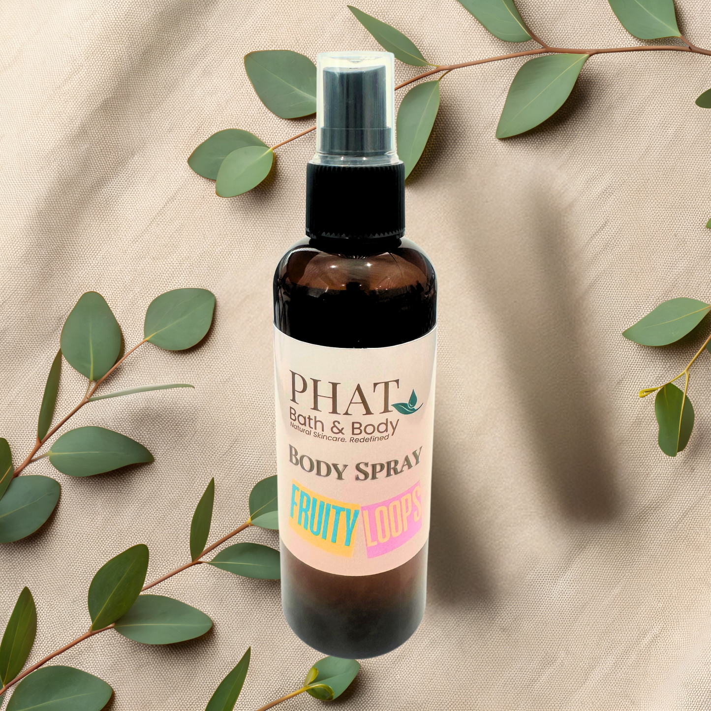 Body spray bottle with 'Phat Bath & Body' label on a beige fabric background with green leaves.