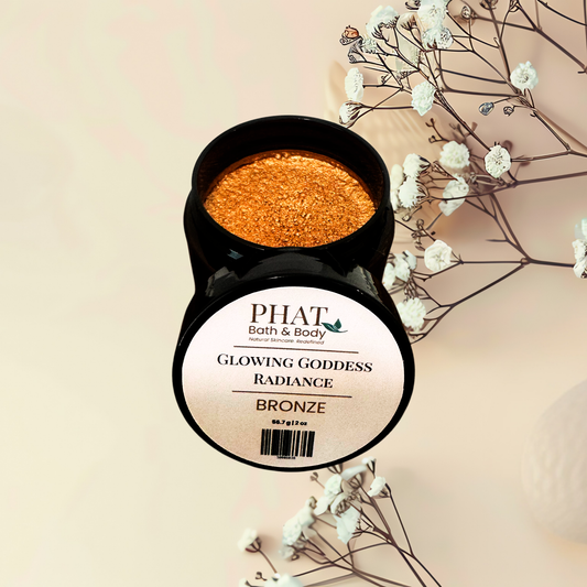 An open black jar of PHAT Bath & Body 'Glowing Goddess Radiance' body powder in the shade 'Bronze', showing the sparkling orange-bronze product inside, styled with dried baby's breath flowers on a warm beige background.