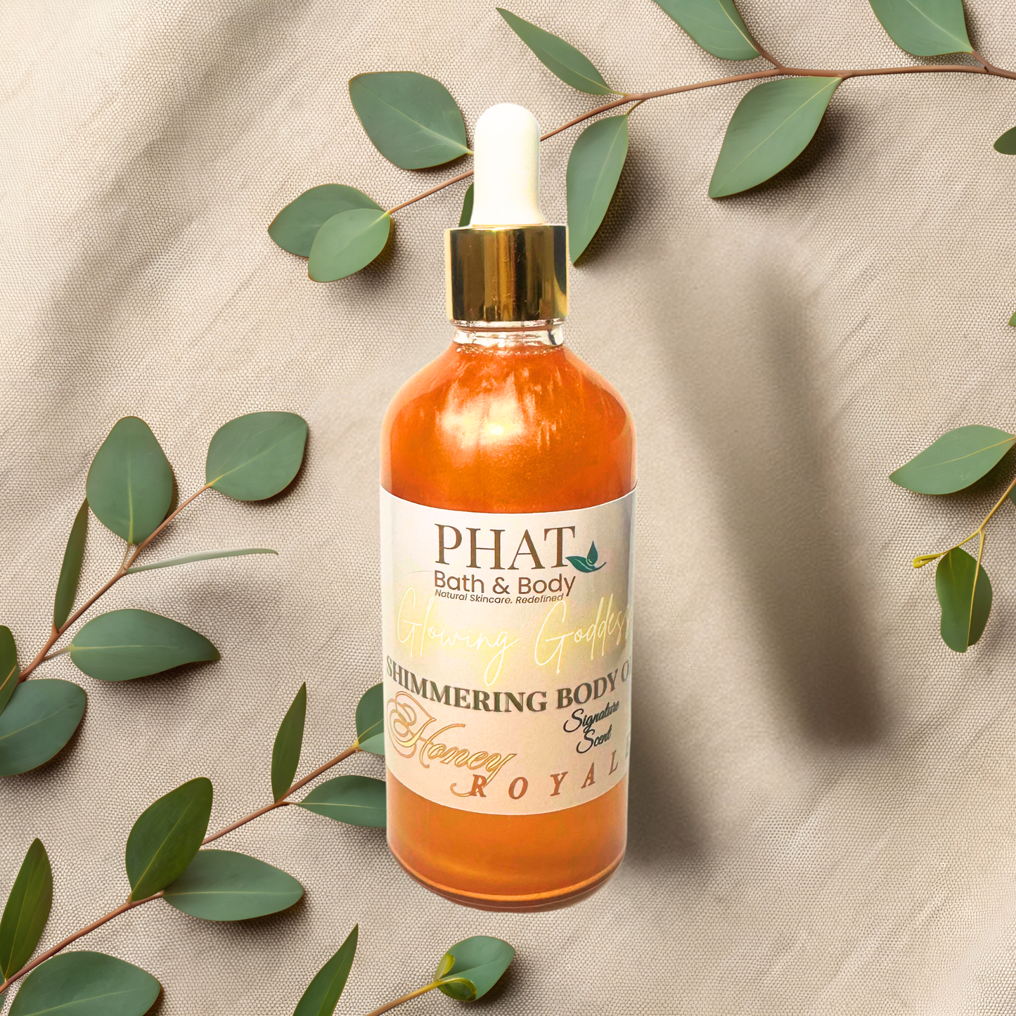 Bottle of 'PHAT Bath & Body' shimmering body oil with leaves on a textured fabric background