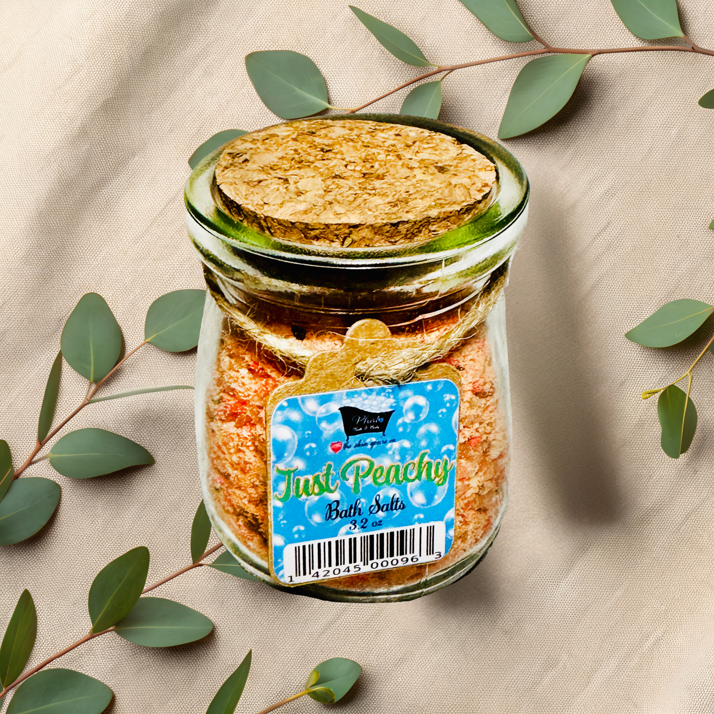 Jar of 'Just Peachy' bath salts with a cork lid on a beige fabric background with green leaves.
