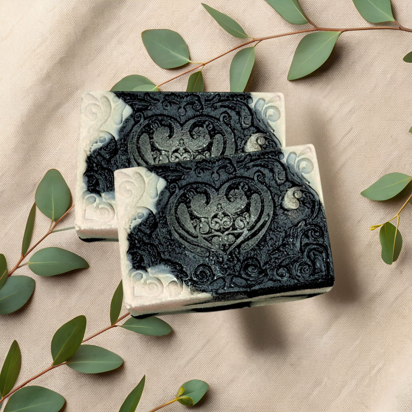 Two bars of black and white soap with intricate designs on a beige fabric background with green leaves.