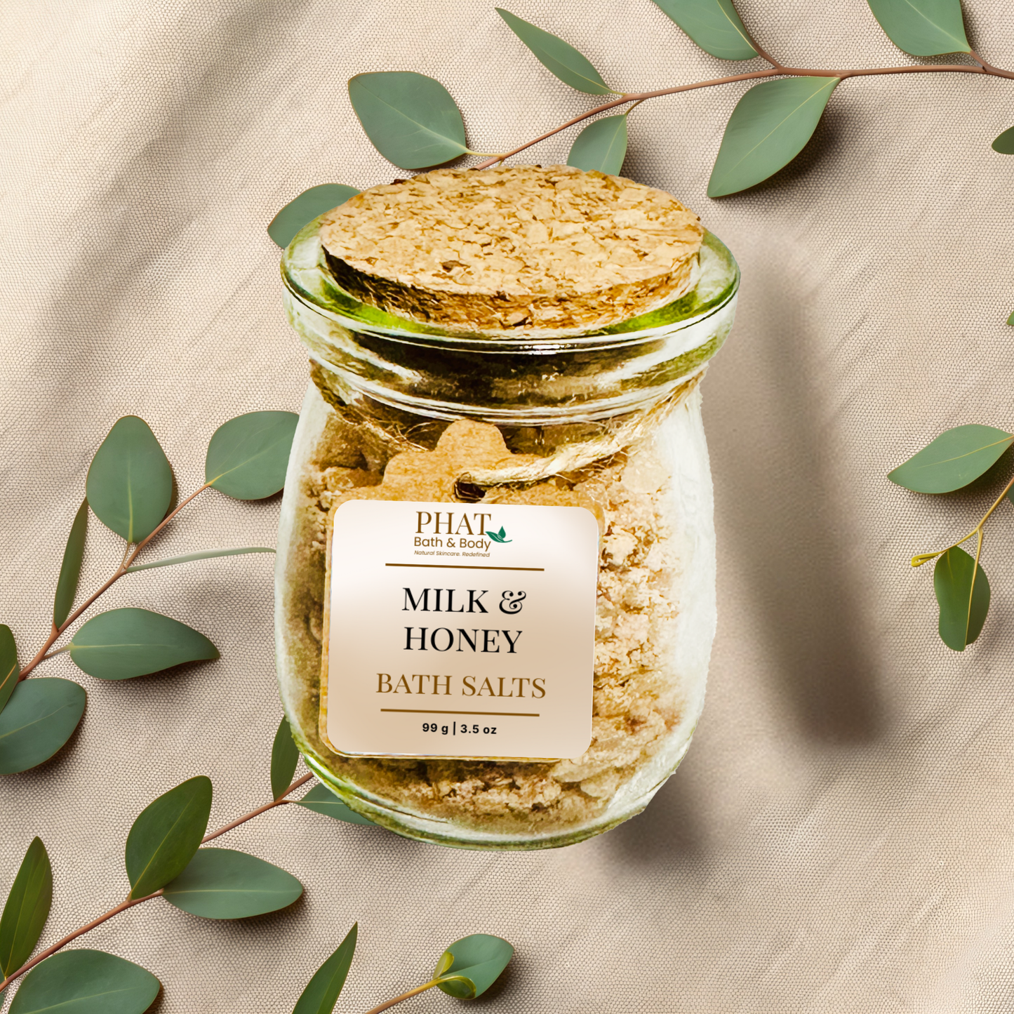 Glass jar of milk and honey bath salts with a cork lid on a beige fabric background with eucalyptus leaves.