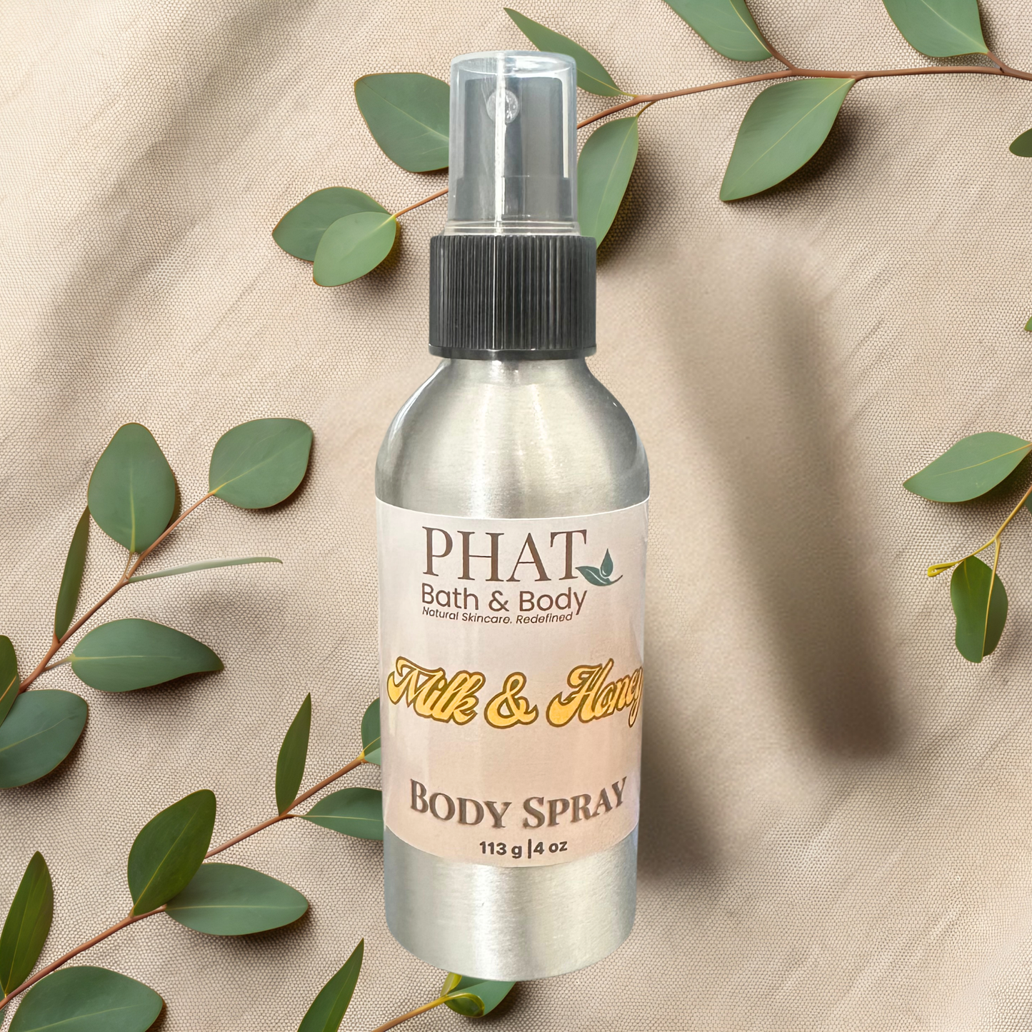 Body spray bottle with 'PHAT Bath & Body' label on a beige fabric background with green leaves.