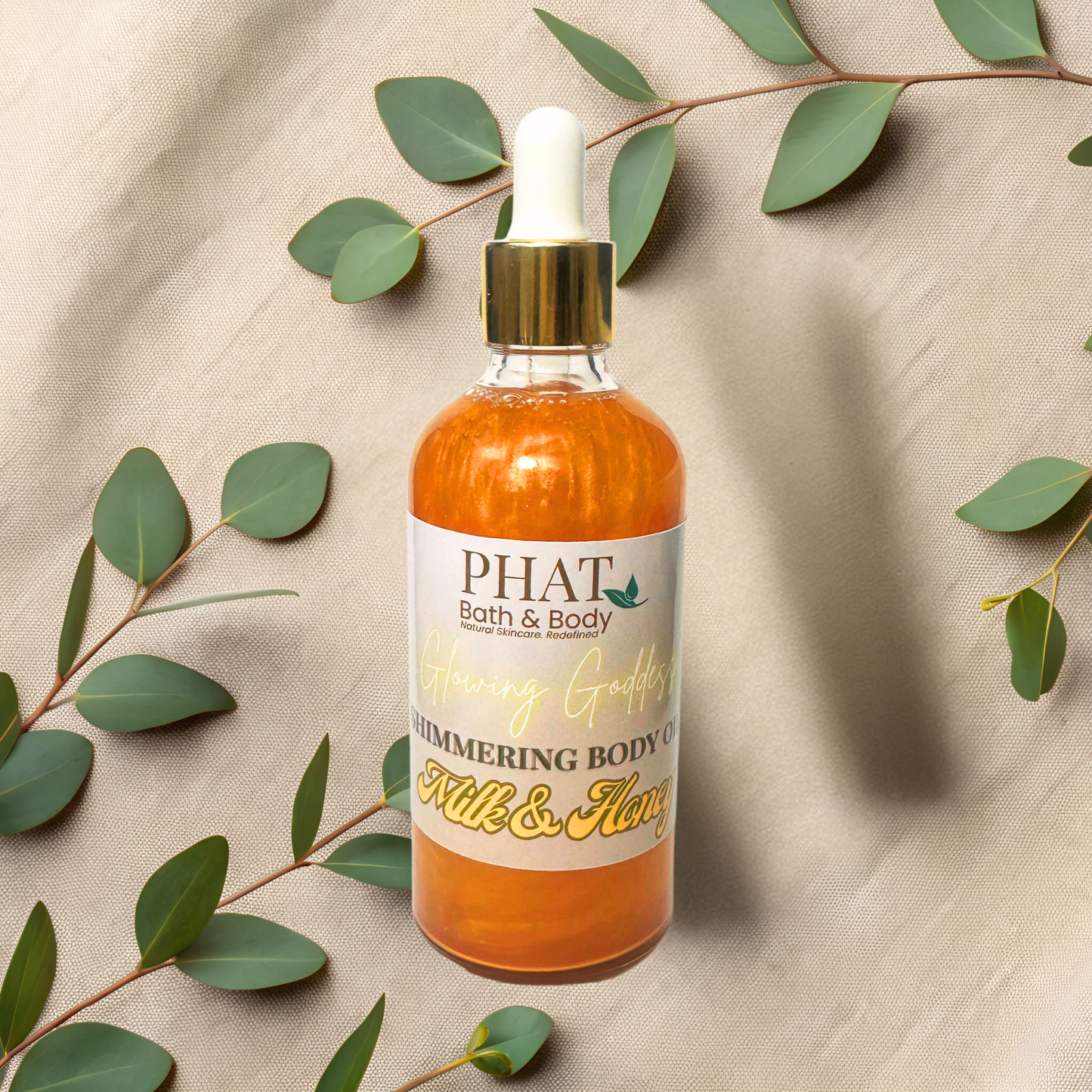 Bottle of 'PHAT Bath & Body' shimmering body oil with green leaves on a beige fabric background