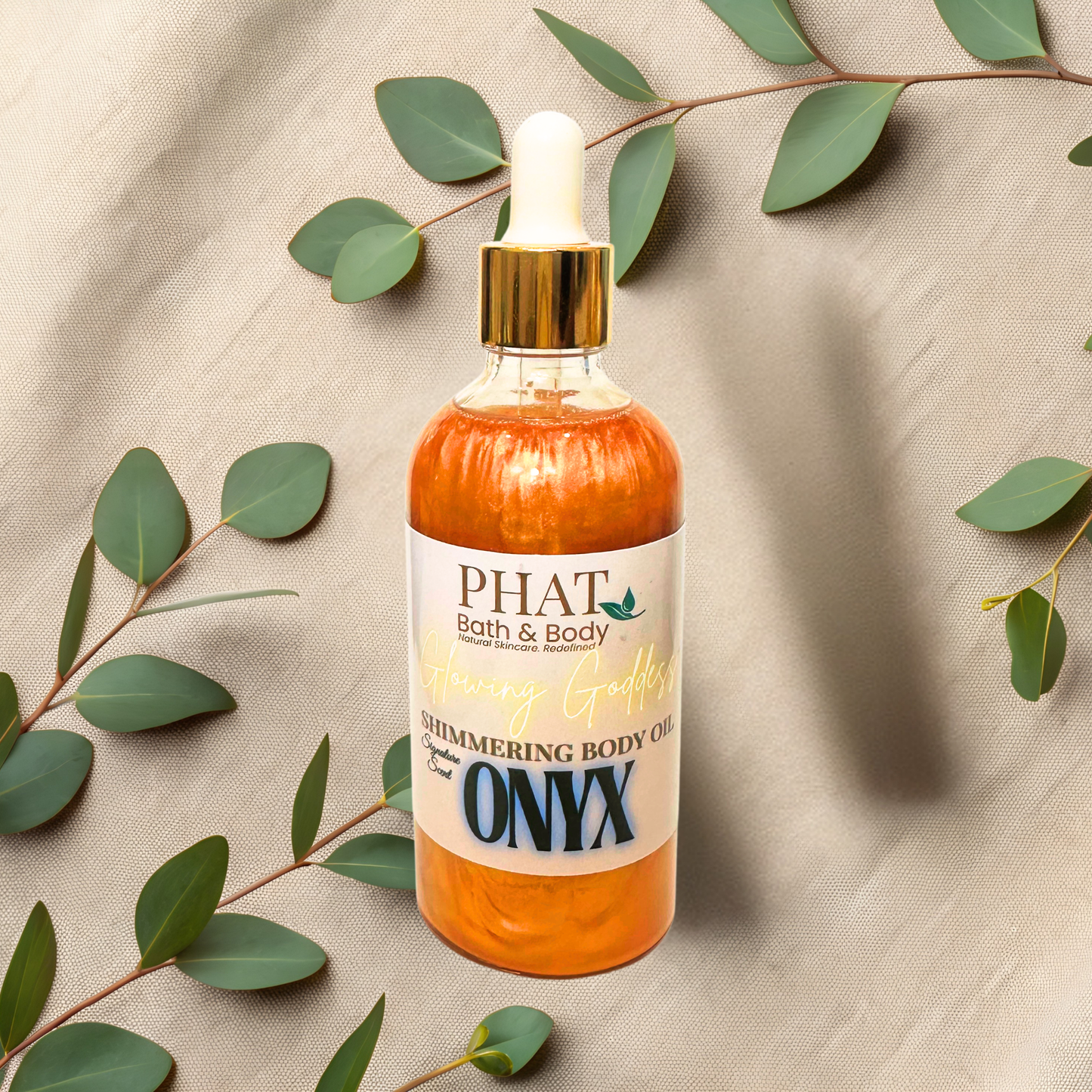 Bottle of 'Onyx' shimmering body oil from PHAT Bath & Body on a beige fabric background with green leaves.