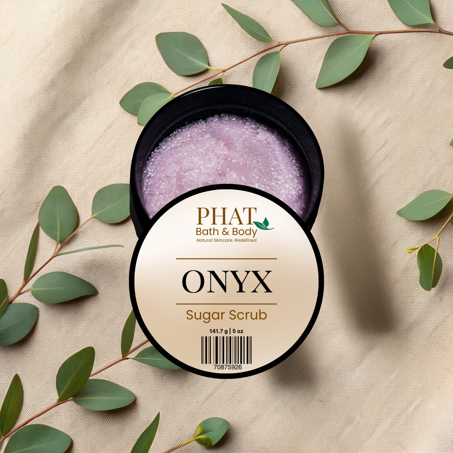 Body scrub jar labeled 'Onyx' by PHAT Bath & Body on a beige fabric background with green leaves.