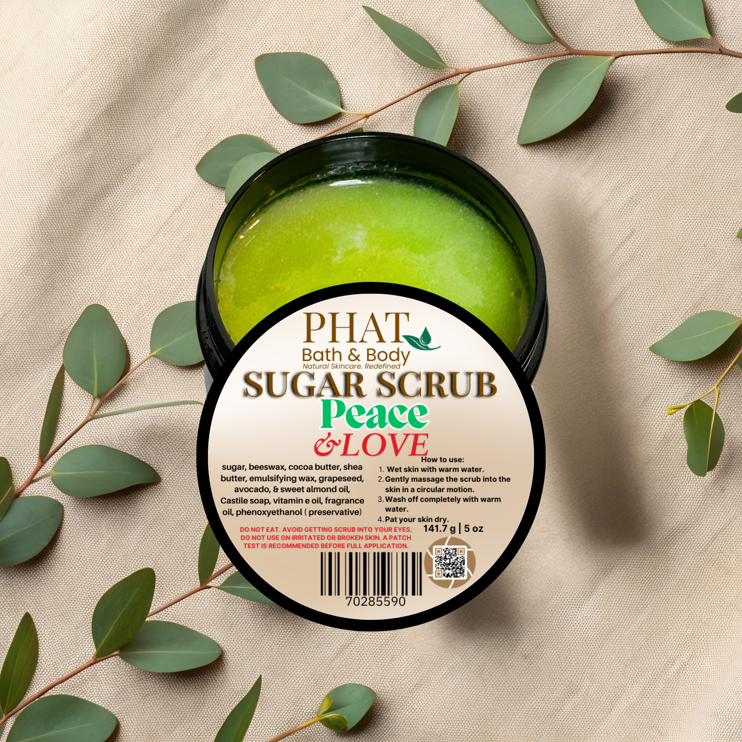 Phat Bath & Body sugar scrub container with green scrub inside, surrounded by eucalyptus leaves on a beige fabric background.