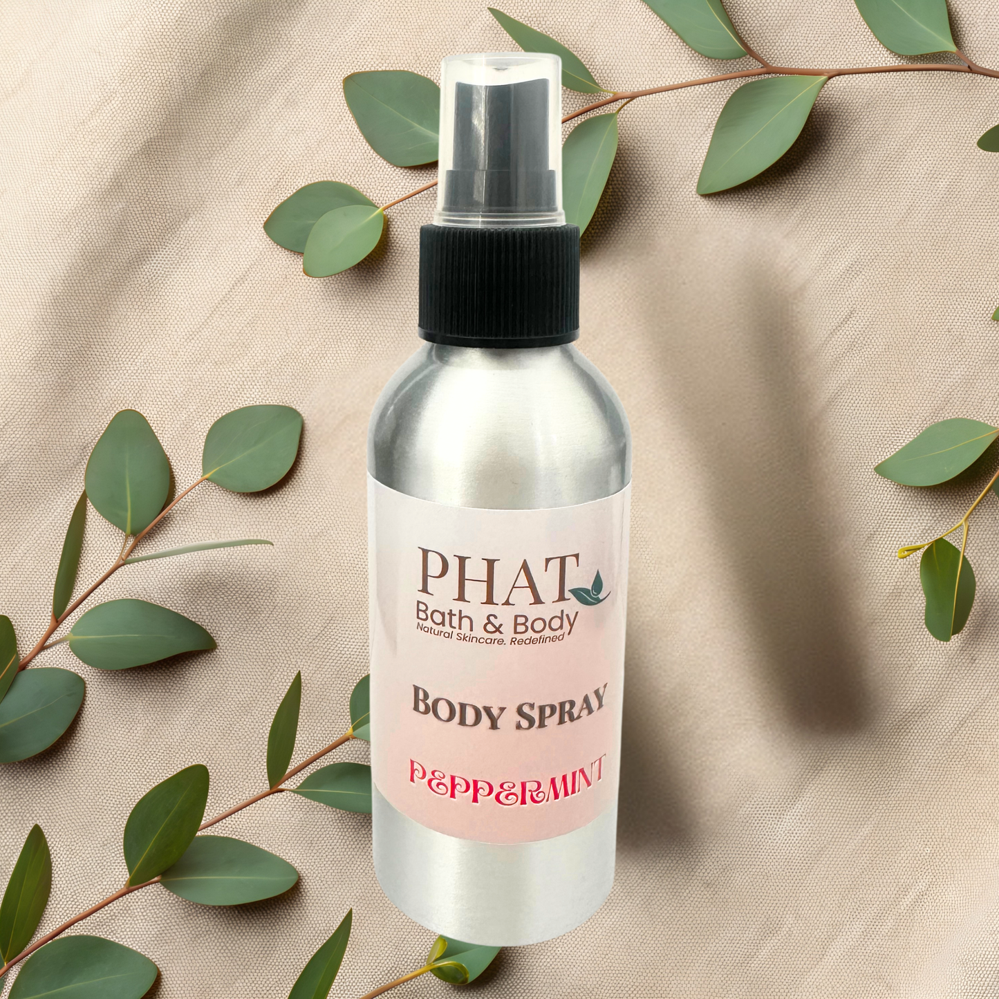 Body spray bottle labeled 'PHAT Bath & Body' with peppermint flavor on a beige fabric background with green leaves.