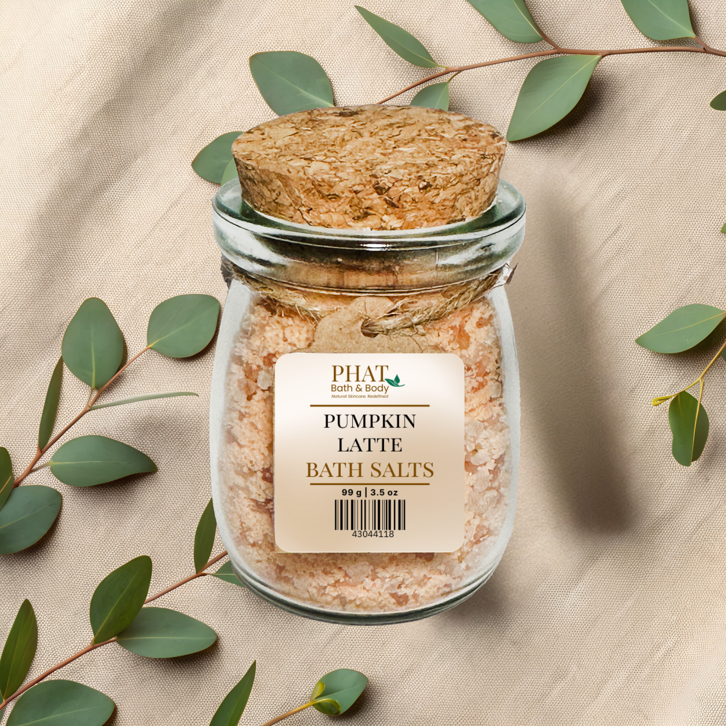 Jar of 'Pumpkin Latte Bath Salts' with a cork lid on a beige fabric background with eucalyptus leaves.