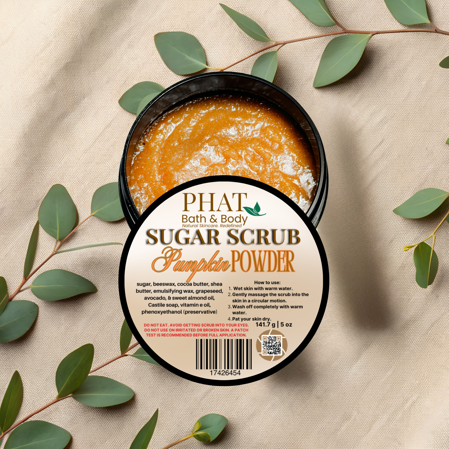 Pumpkin powder sugar scrub in a container with a 'Phat Bath & Body' label on a beige fabric background with green leaves.
