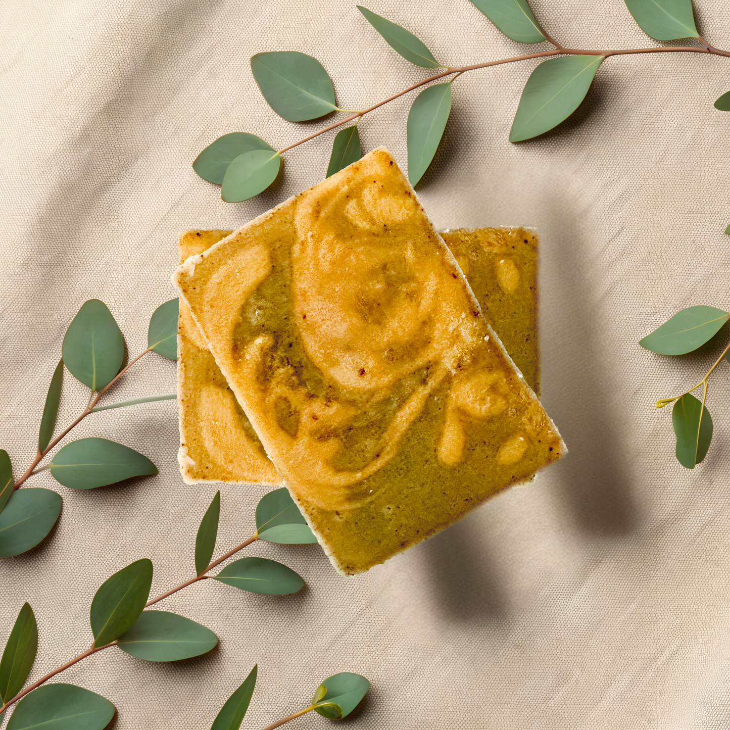 Two square yellowish-green bars of soap on a beige fabric background with green leaves.