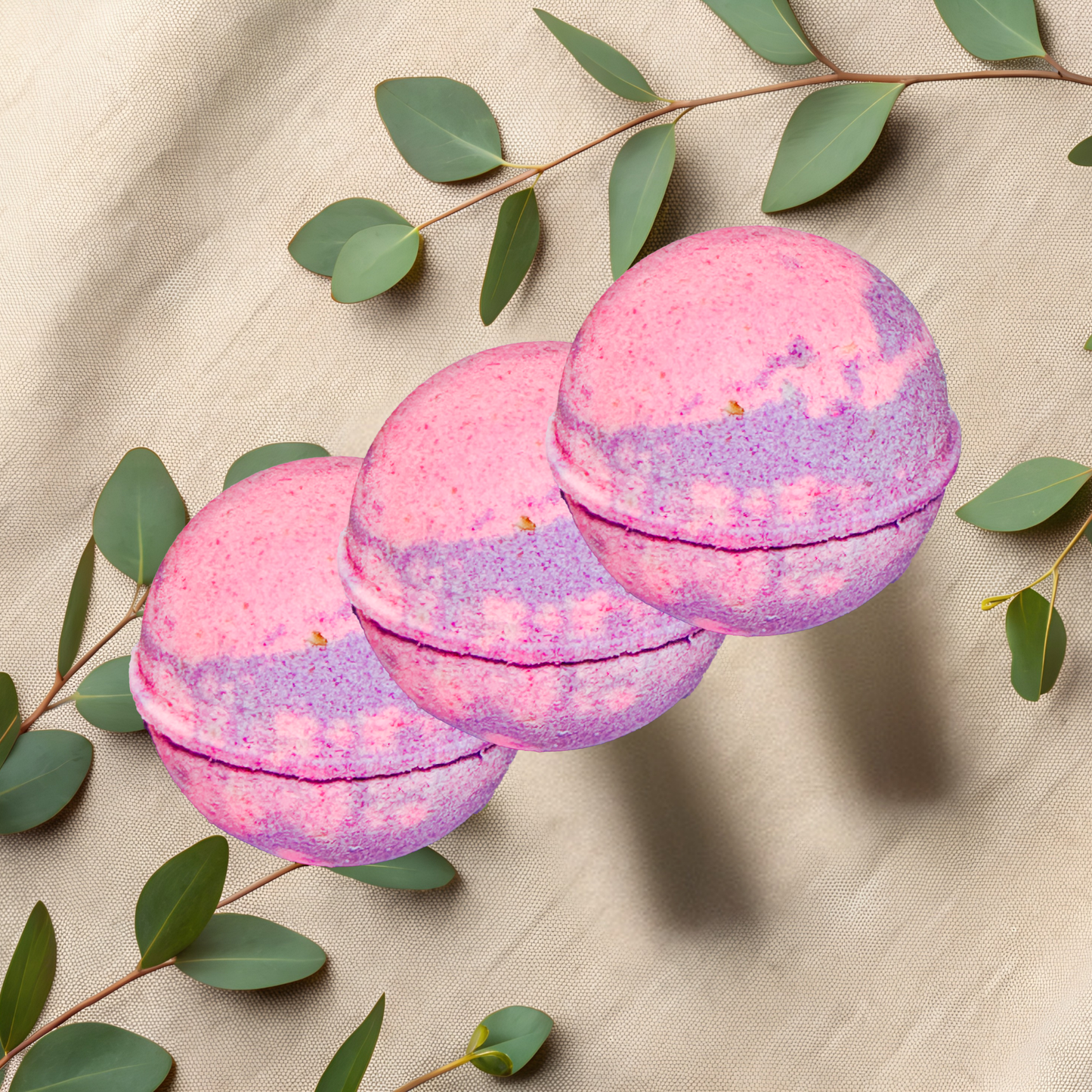 Three pink and purple bath bombs with green leaves on a beige fabric background