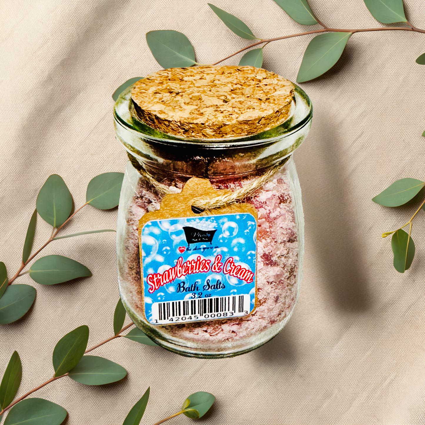 Jar of 'Strawberries & Cream' bath salts with a cork lid on a beige fabric background with green leaves.