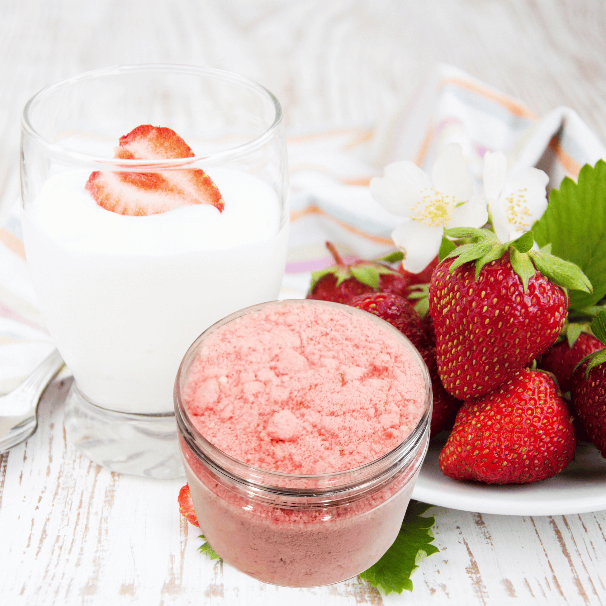 Jar of strawberry-scented bath bomb powder, glass with a strawberry slice, and fresh strawberries on a white surface.