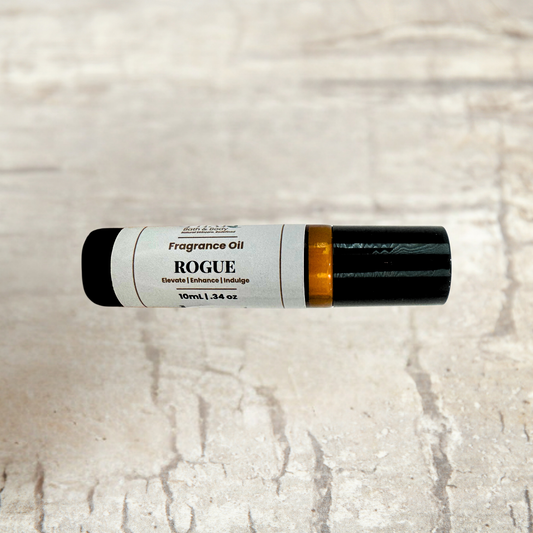 A 10ml tube of Rogue Fragrance Oil decant fragrance, featuring a white label with black text and a black cap, resting horizontally on a light beige stone-textured background.