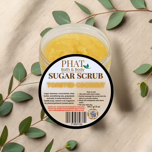 Jar of Phat Bath & Body Sugar Scrub with toasted coconut flavor on a beige fabric background with green leaves.