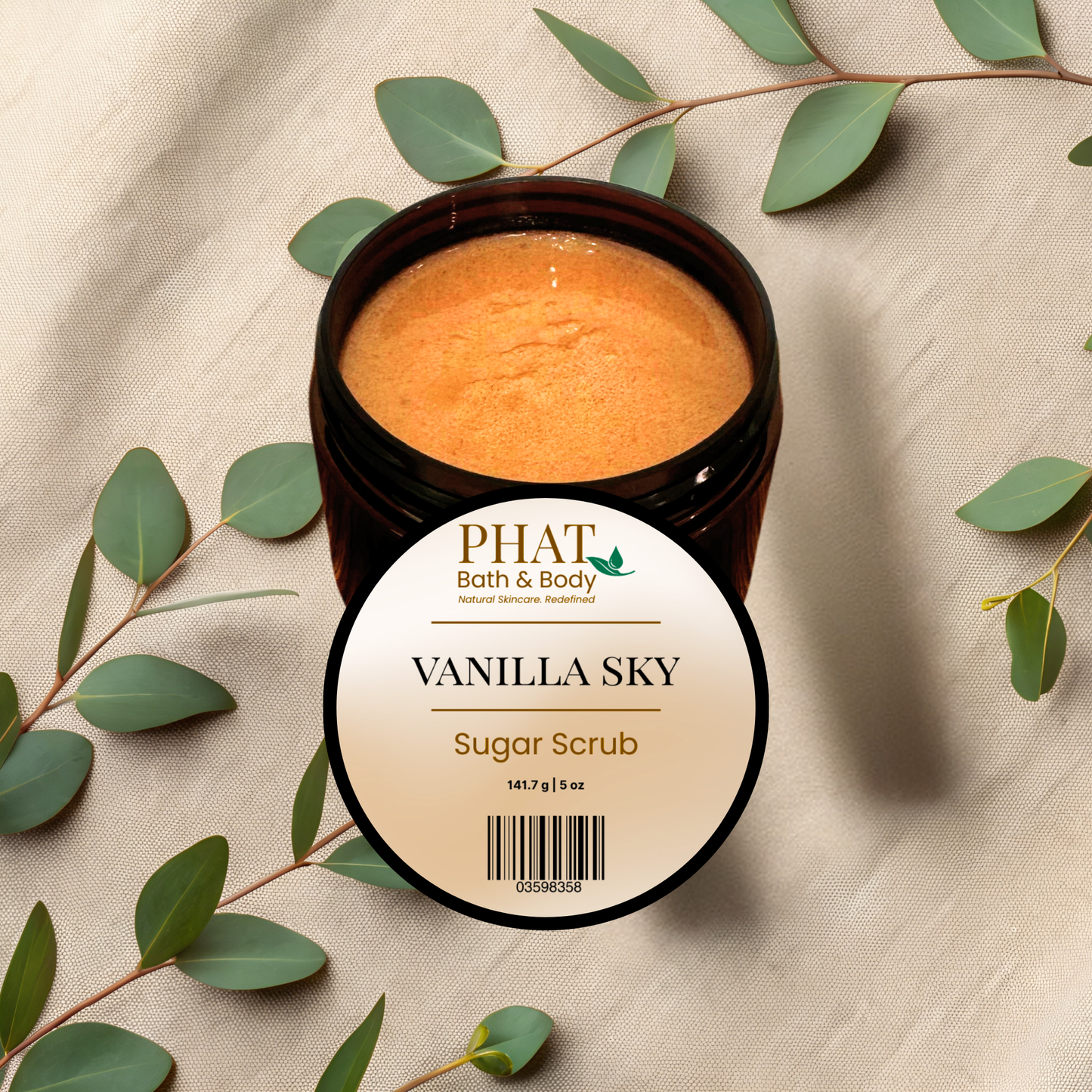 An open dark amber jar of PHAT Bath & Body Vanilla Sky Sugar Scrub, showing the brown scrub inside, styled on a beige fabric background with eucalyptus leaves.