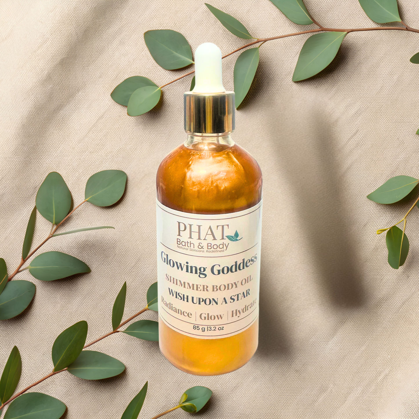 Bottle of PHAT Bath & Body Glowing Goddess Shimmer Body Oil on a beige fabric background with green leaves.