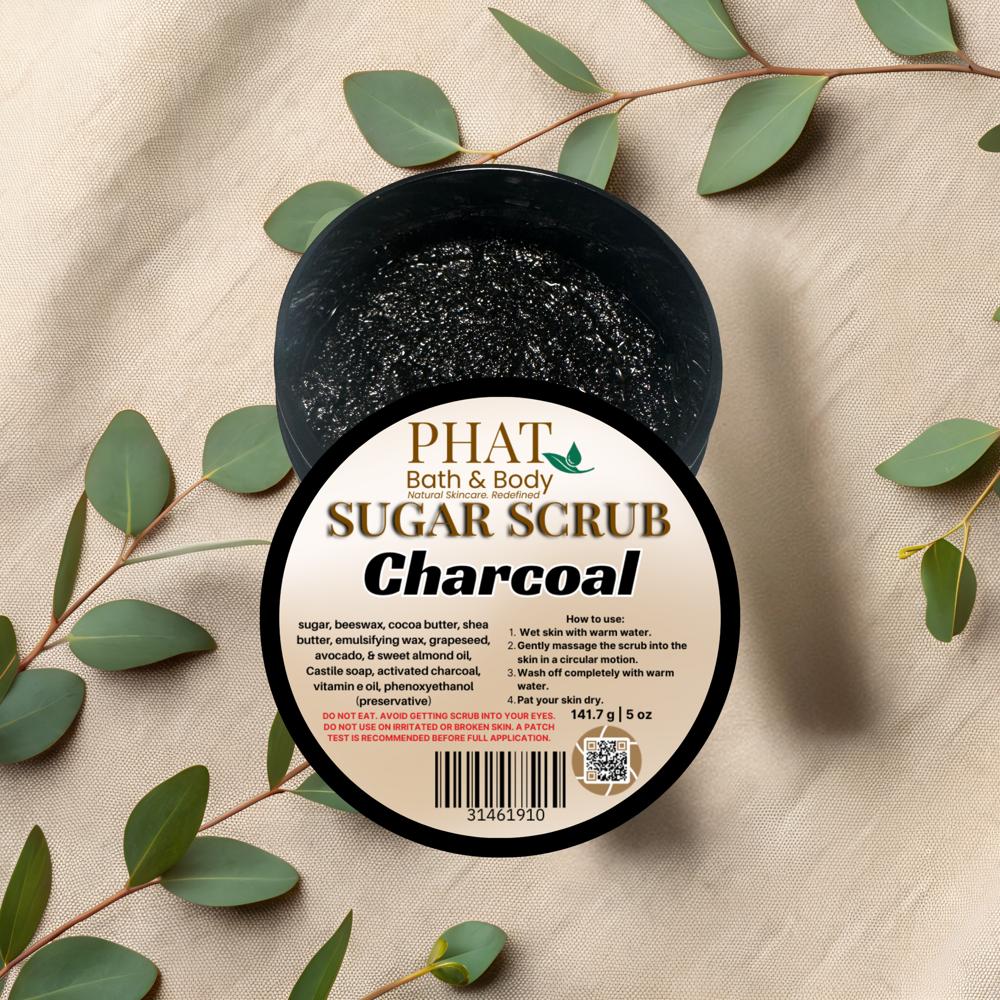 Jar of charcoal sugar scrub with a label on a beige fabric background with green leaves.