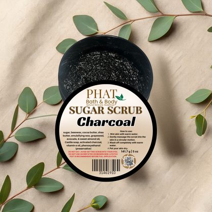 Jar of charcoal sugar scrub with a label on a beige fabric background with green leaves.