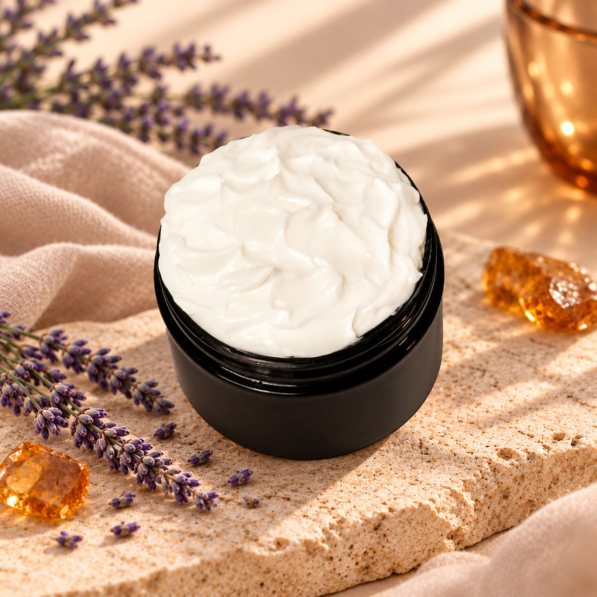 Open jar of Amber & Lavender Body Lotion styled with lavender sprigs, warm amber accents, soft fabric, and light natural stone.