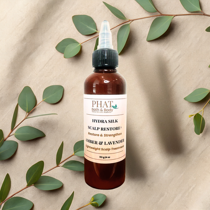 An amber bottle of PHAT Bath & Body Hydra Silk Scalp Restore treatment, styled on a beige textured fabric background with eucalyptus leaves.