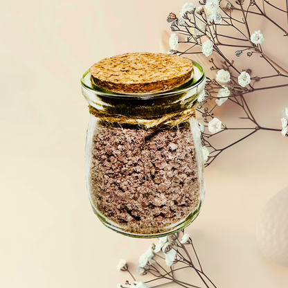Glass jar with cork lid containing a brown bath salts, surrounded by white flowers on a beige background