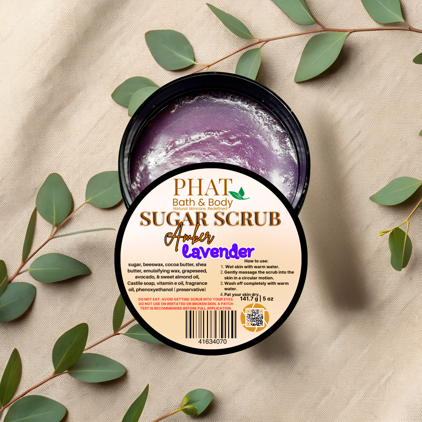 PHAT Bath & Body Sugar Scrub container with purple scrub on a beige fabric background with green leaves.