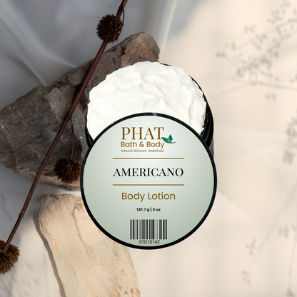 A round tub of PHAT Bath & Body 'Americano' body lotion, showing the white cream inside, styled with natural wood pieces and dried brown seed pods on a light background.