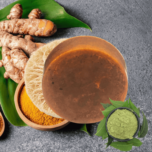 Brown soap with turmeric powder, fresh turmeric roots, and green leaves on a gray surface