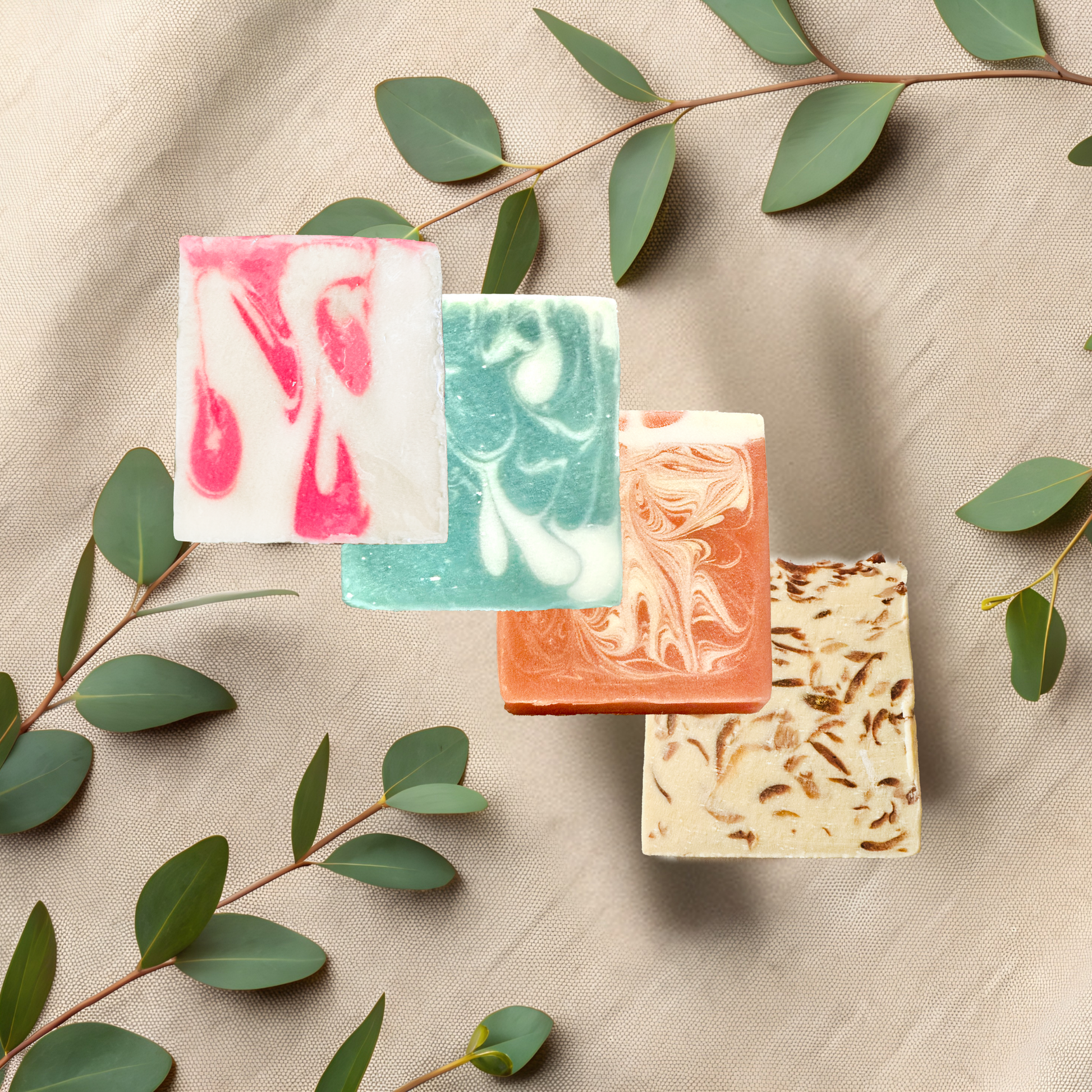 Four different colored soap bars on a beige fabric background with green leaves.