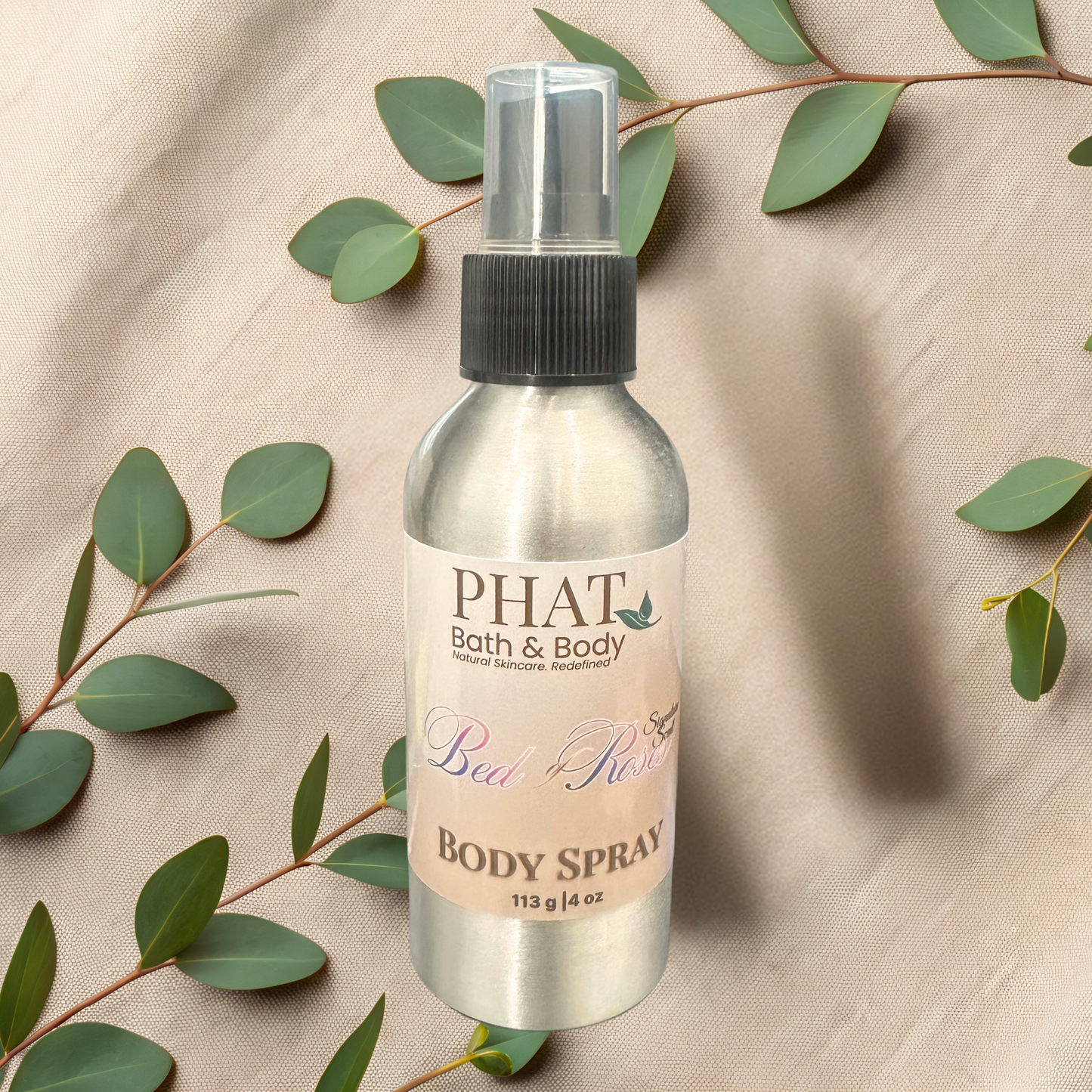 Body spray bottle with 'PHAT Bath & Body' label on a beige fabric background with green leaves.