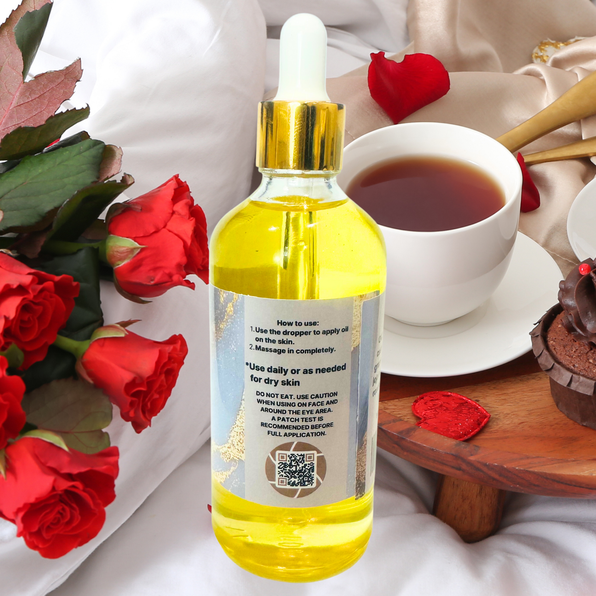 Bottle of yellow body oil with dropper, surrounded by red roses and a cup of tea on a white surface.