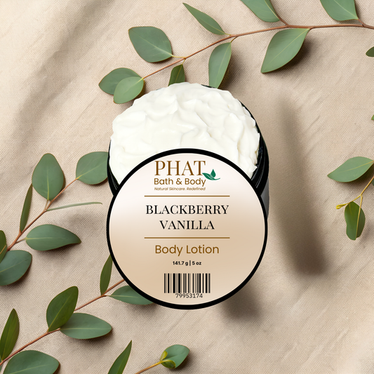 A round-labeled tub of PHAT Bath & Body Blackberry Vanilla Body Lotion filled with whipped white lotion, styled on a beige fabric background with eucalyptus leaves.