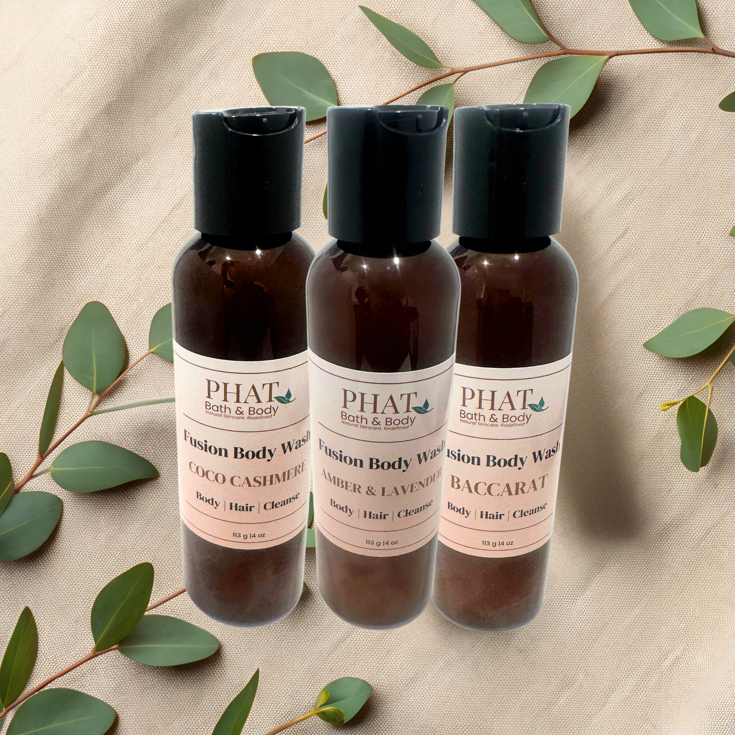 Three bottles of PHAT Bath & Body Fusion Body Wash on a beige fabric background with green leaves.