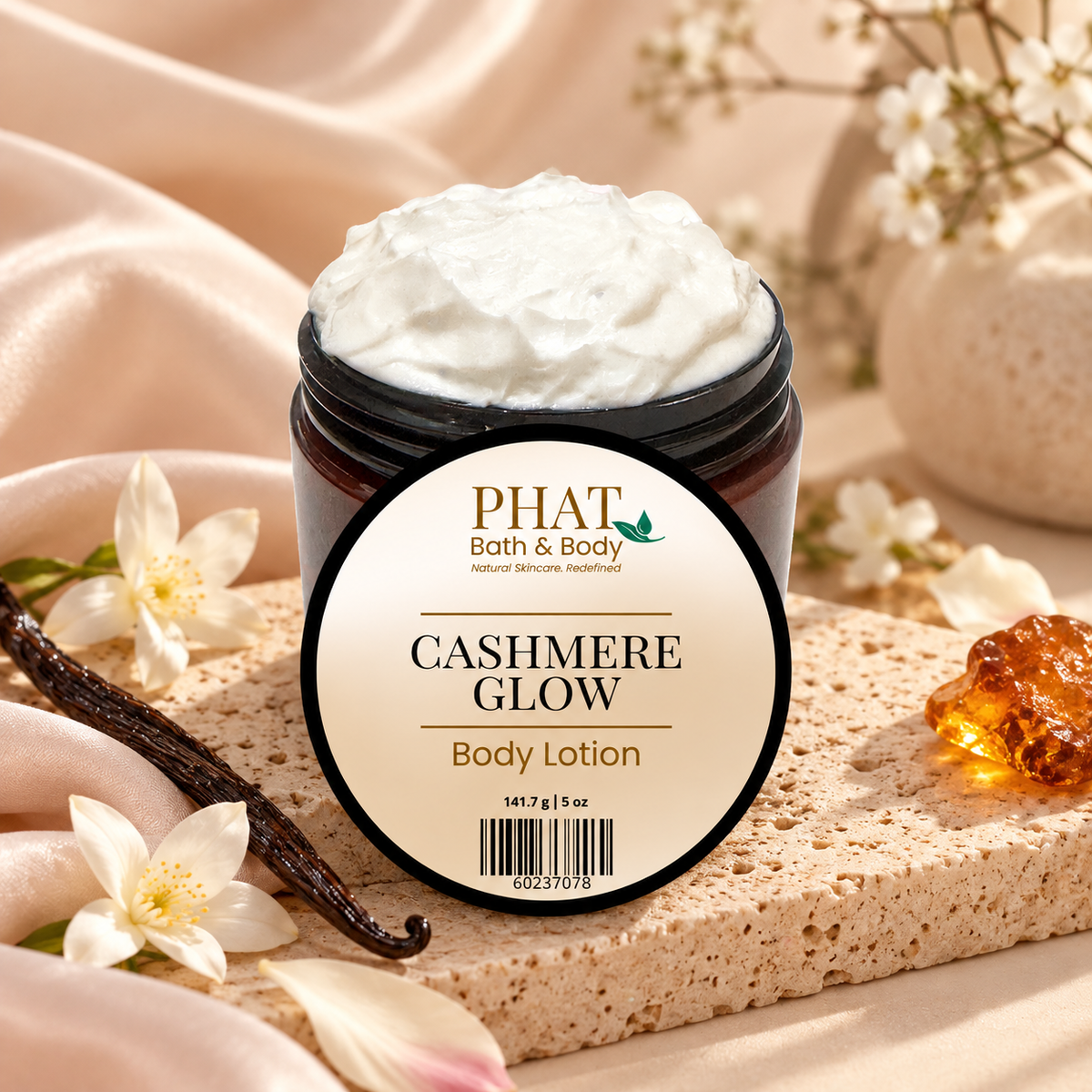 Phat Bath & Body Cashmere Glow 5 oz Body Lotion in an open black jar with labeled lid, styled with vanilla beans, white florals, warm amber accents, and soft neutral fabric.