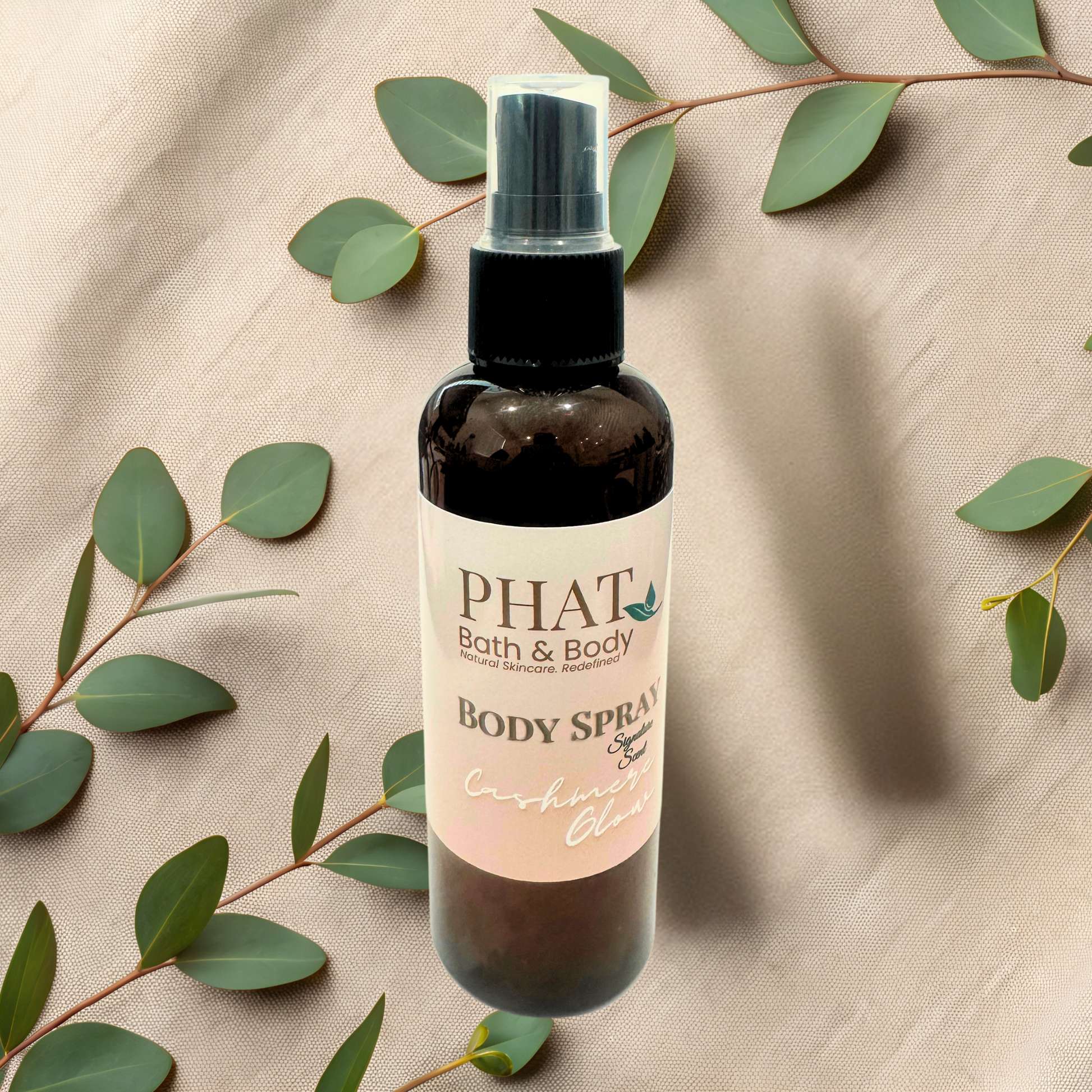 Body spray bottle with 'PHAT Bath & Body' label on a beige fabric background with green leaves.