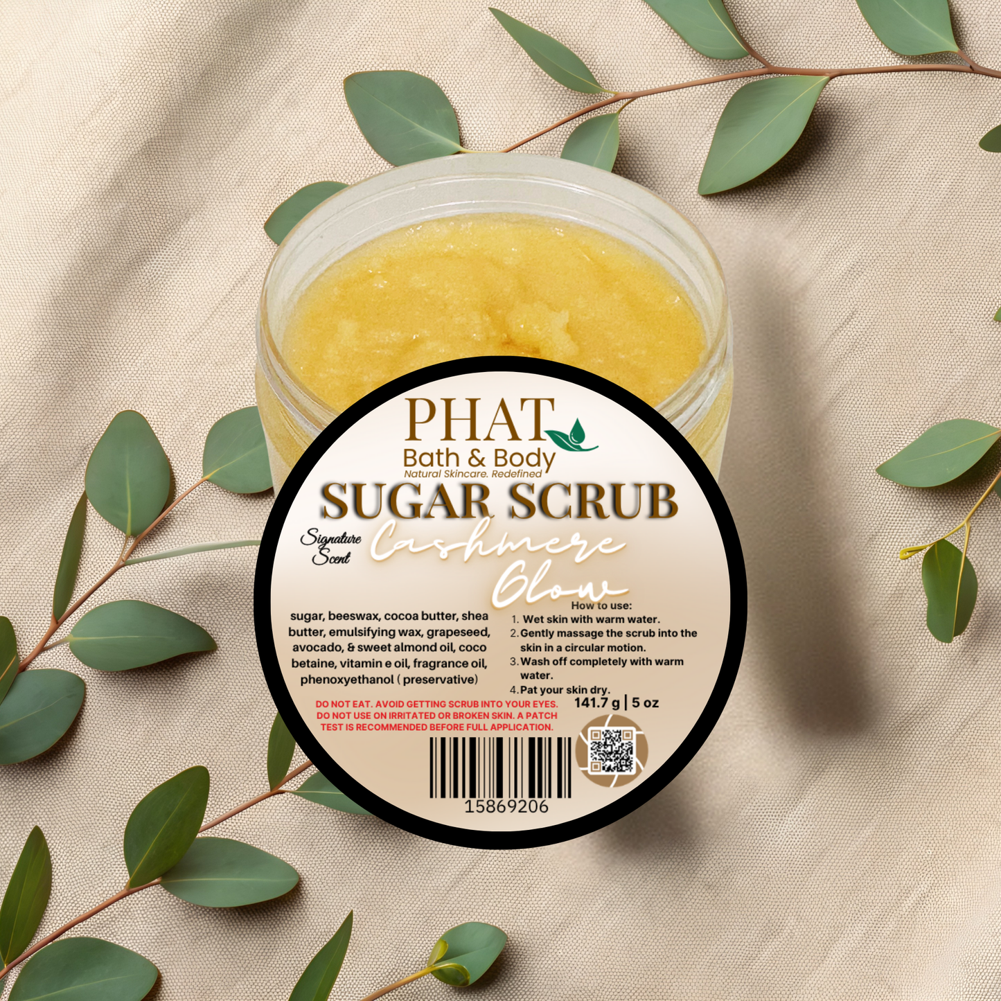 Jar of sugar scrub with a label on a beige fabric background with green leaves.