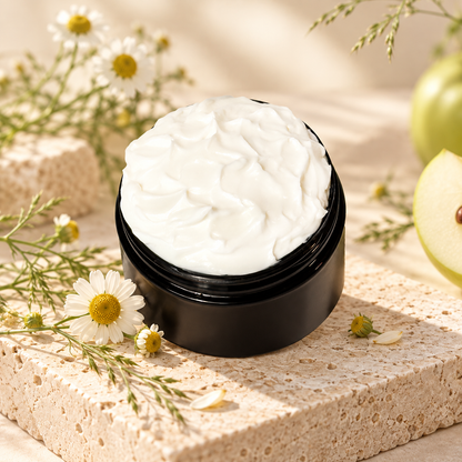 Open jar of Phat Bath & Body Chamomile Body Lotion displayed with chamomile flowers, fresh green apple, delicate greenery, and light neutral stone in a clean botanical setting.