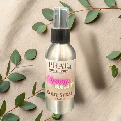 Body spray bottle labeled 'PHAT Bath & Body Cherry Blossom' on a beige fabric background with green leaves.