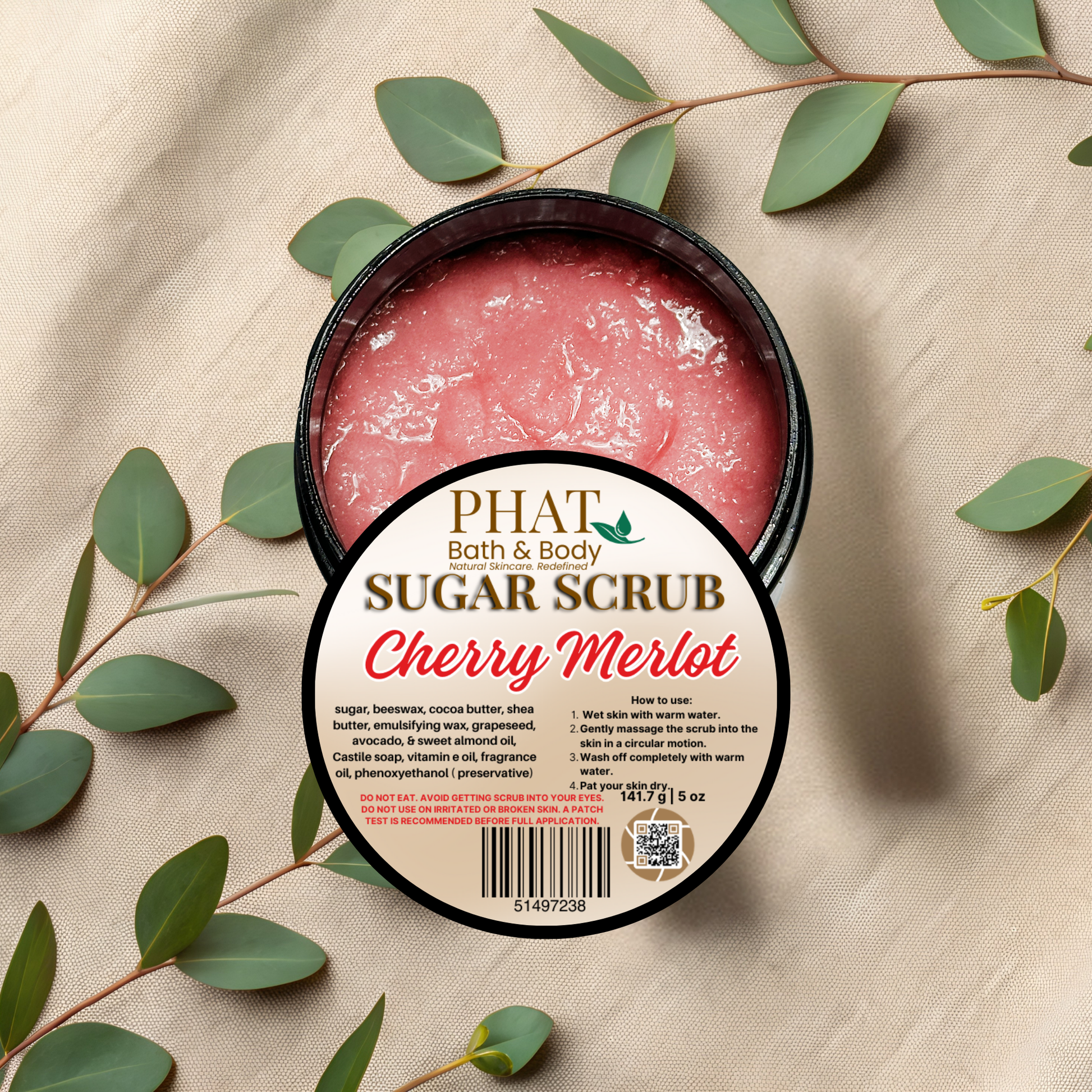 Jar of cherry merlot sugar scrub with a 'Phat Bath & Body' label on a beige fabric background with green leaves.